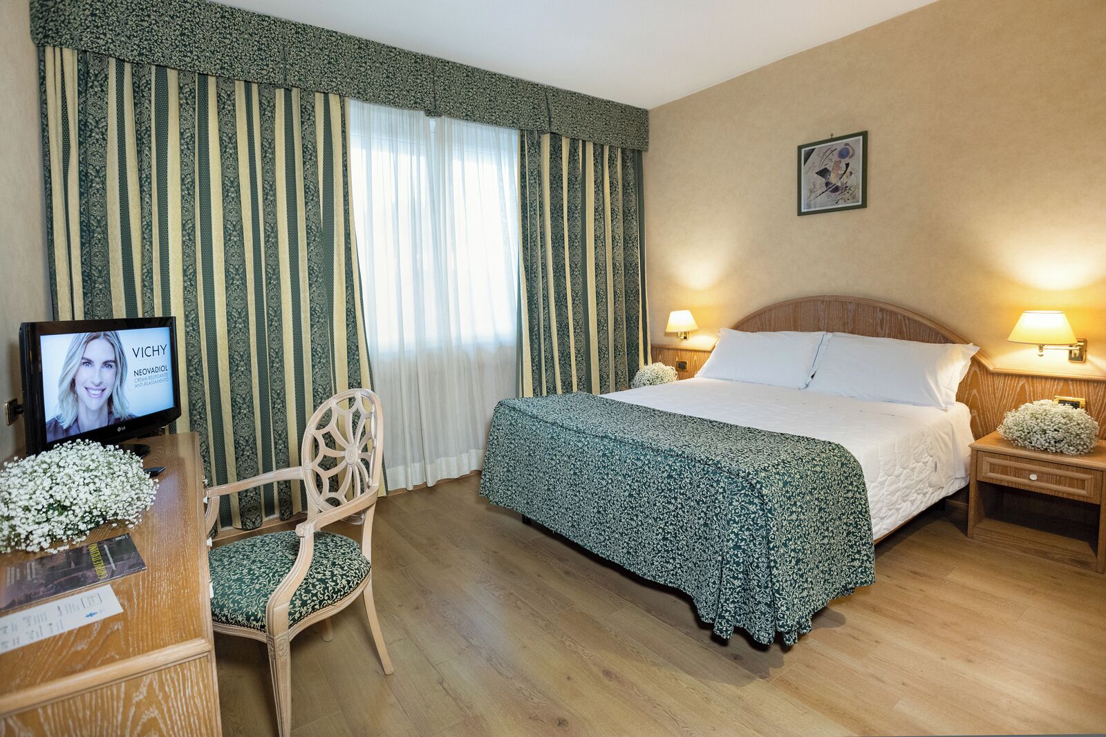 A hotel room with a bed, two bedside tables with lamps, a wooden chair, a television, and a large window with curtains.