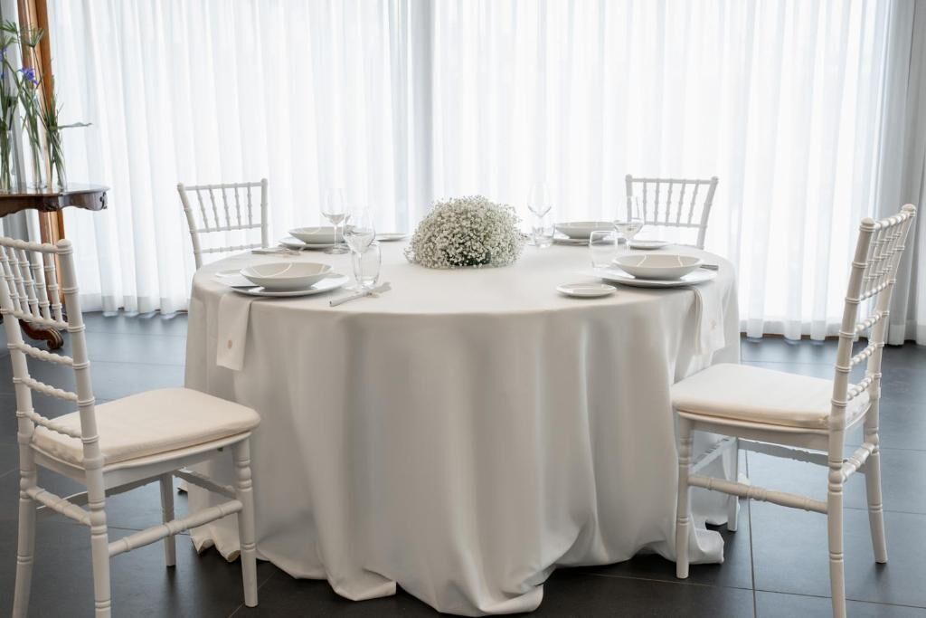 A round dining table with white chairs, a white tablecloth, and a centerpiece.