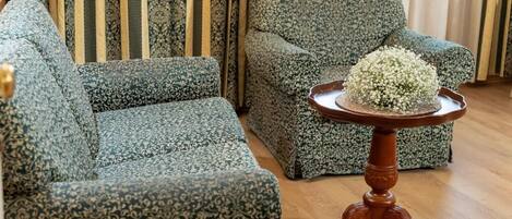 Uma sala de estar com sofás verdes estampados, uma mesa de centro de madeira com um arranjo floral e cortinas listradas.