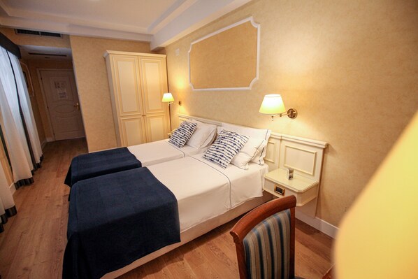 A hotel room with two beds, a wooden floor, a chair, and a wall-mounted lamp.