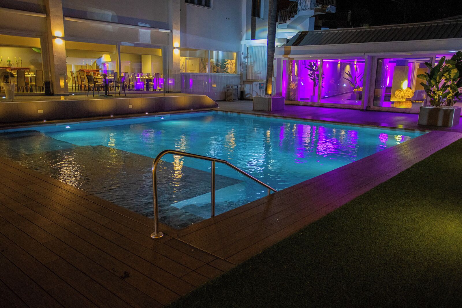 A swimming pool with a wooden deck and a glass barrier, surrounded by a building with purple lighting.
