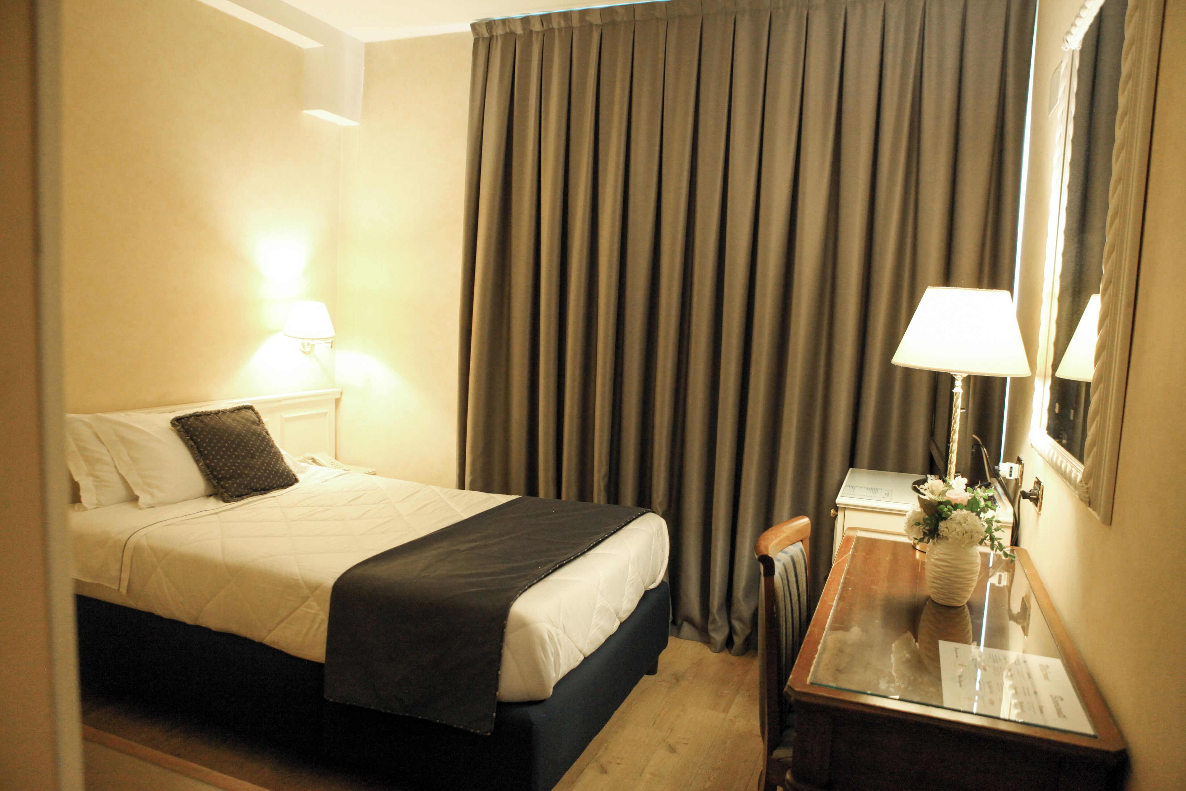 Une chambre d’hôtel avec un lit, un bureau doté d’une lampe, un miroir et une fenêtre avec des rideaux.