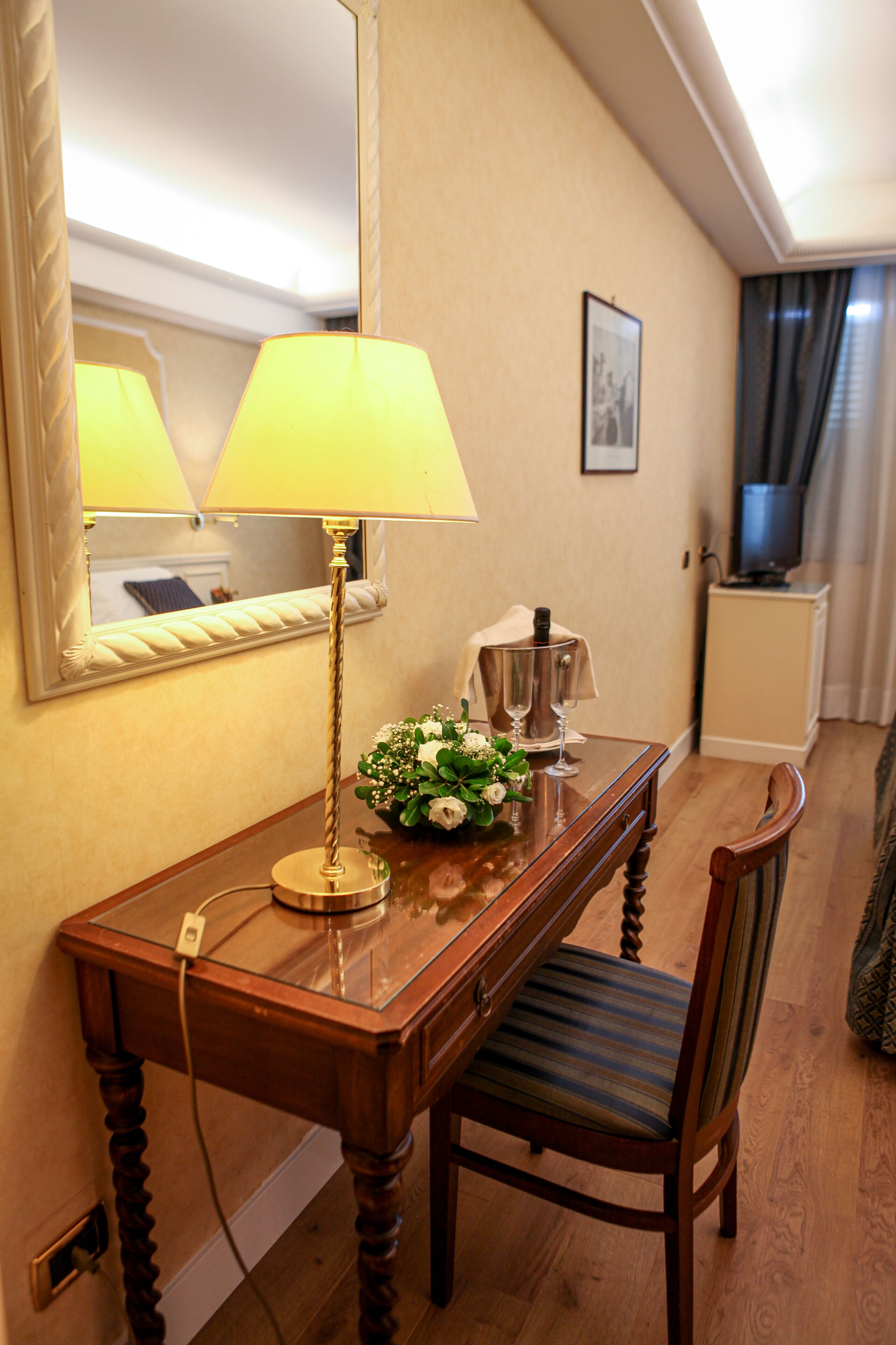 A hotel room with a wooden desk, a lamp, a vase with flowers, a framed picture on the wall, and a television in the corner.