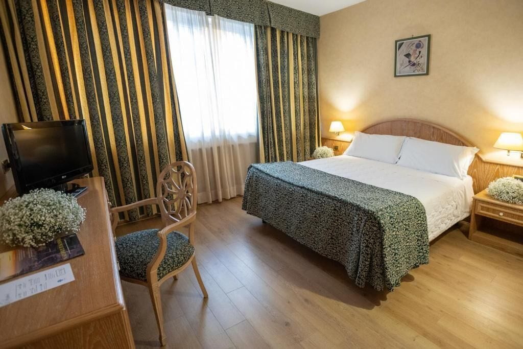 Habitación de hotel con cama, escritorio con televisor, silla y ventana con cortinas.