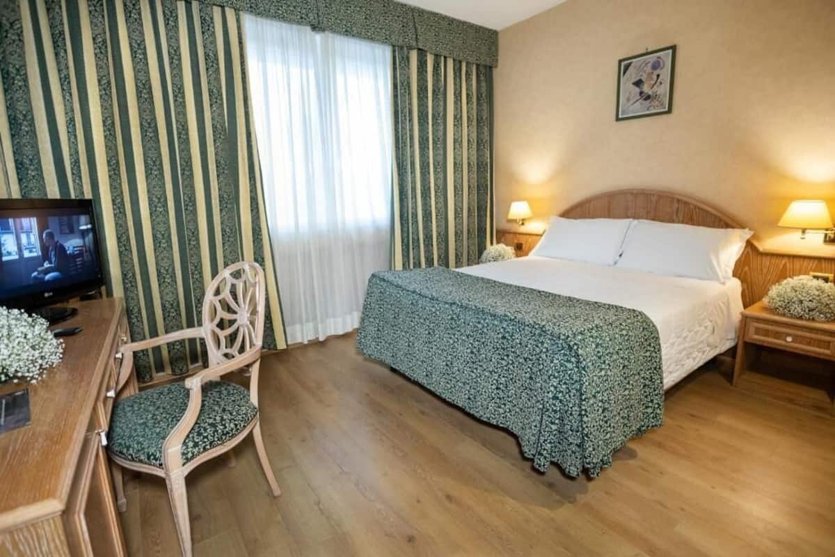 Una habitación de hotel con una cama, dos mesitas de noche con lámparas, un escritorio de madera con una silla, un televisor y una ventana con cortinas a rayas.