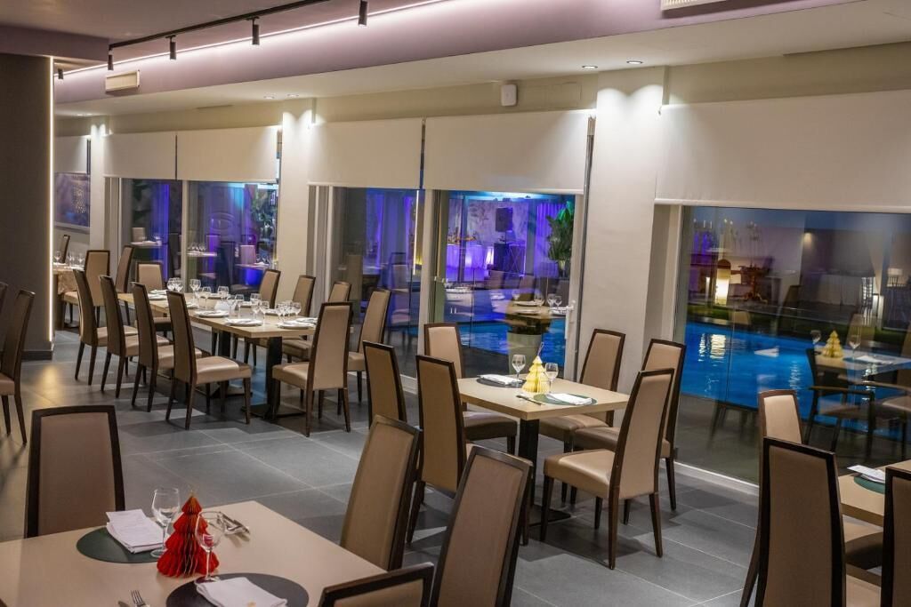 Un espace repas moderne, avec des tables et des chaises, donnant sur une piscine, et une cloison en verre séparant l’espace repas d’un autre espace.