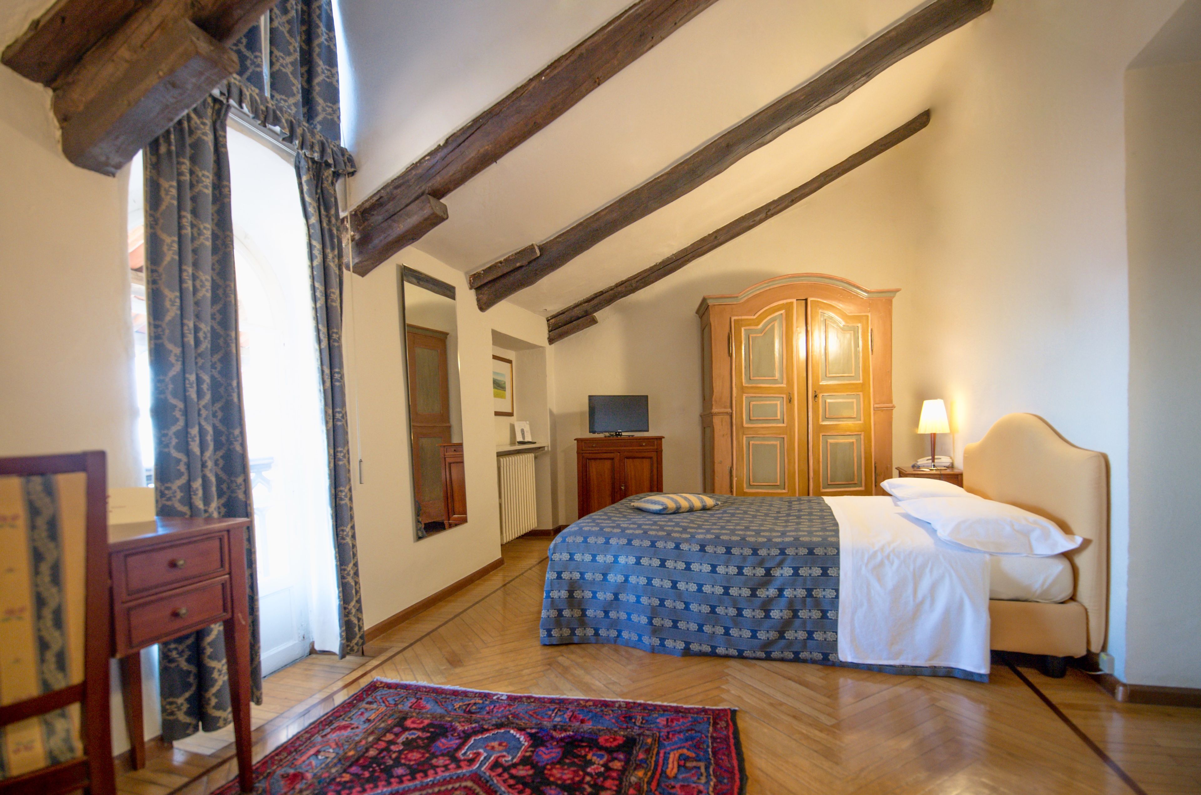A bedroom with a wooden ceiling, a bed with a patterned bedspread, a wooden wardrobe, a television, and a bedside table with a lamp.