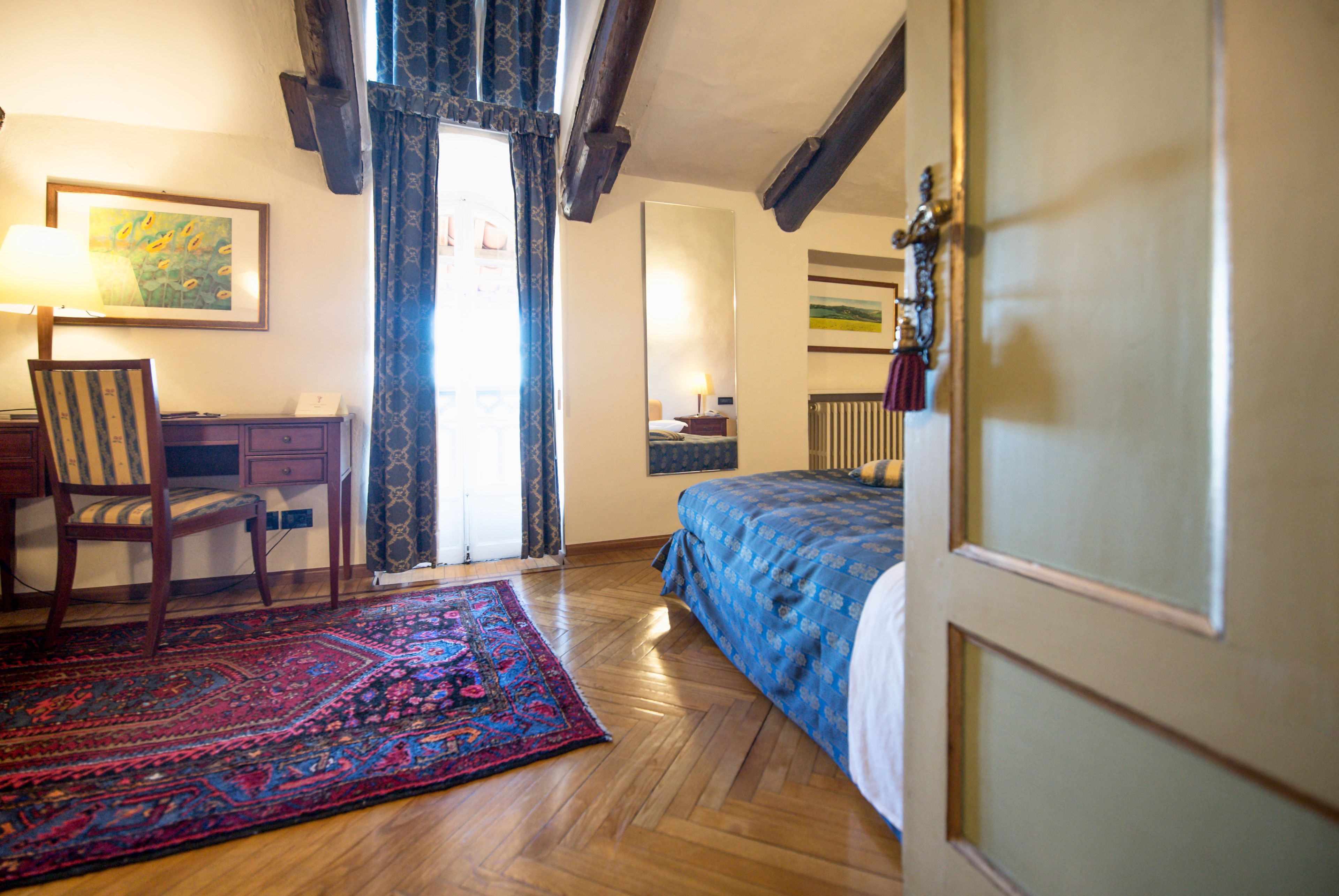 A bedroom with a wooden floor, a bed with a blue bedspread, a chair, a desk, a mirror, and a window with curtains.
