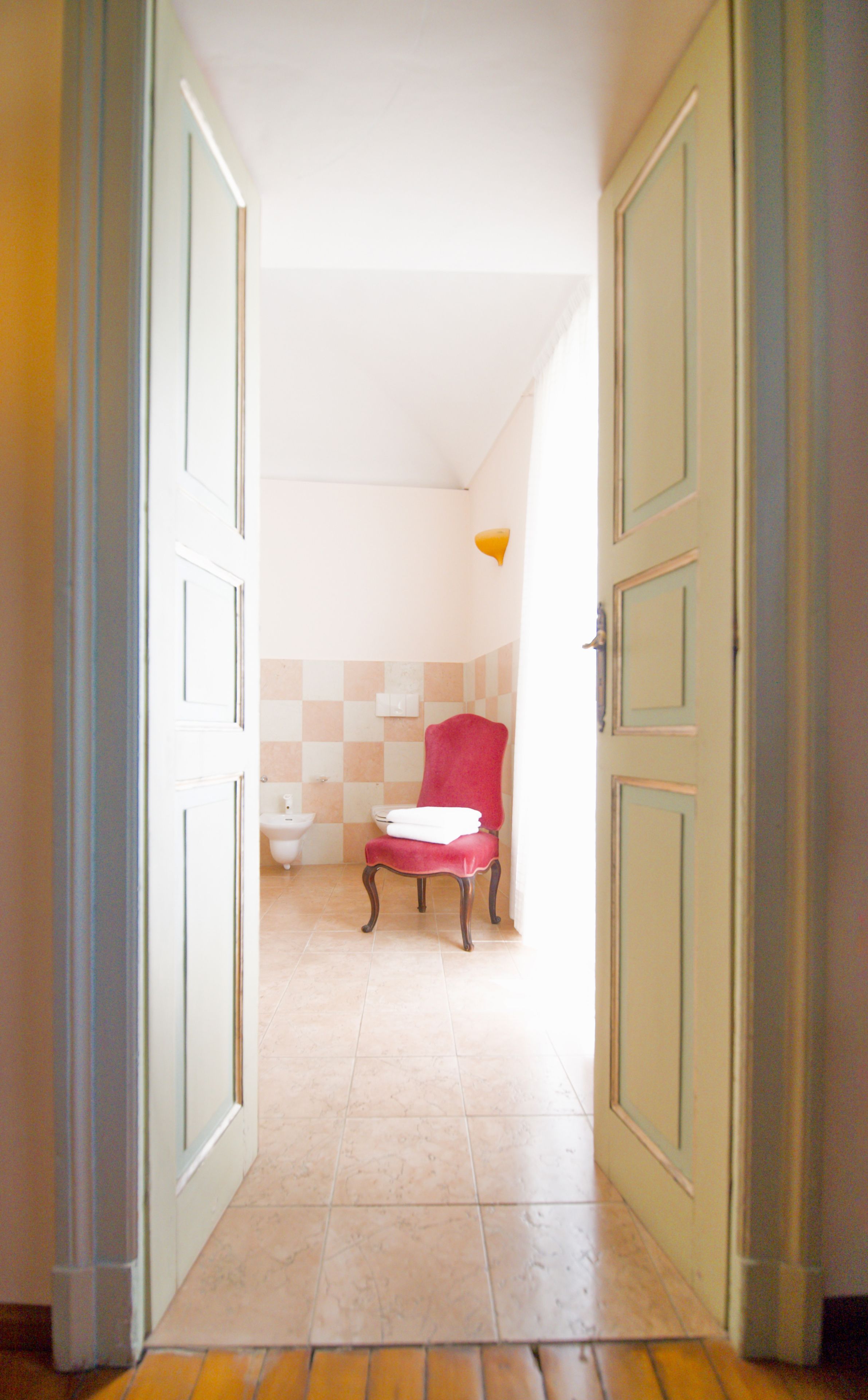 A hallway leading to a bathroom with a red chair, a toilet, and a light fixture.