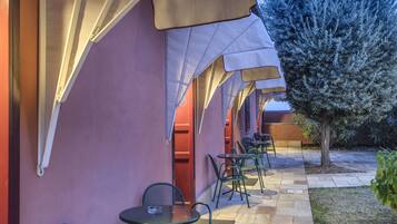 An outdoor seating area with tables and chairs, a tree, and a building with a red wall and white awnings.