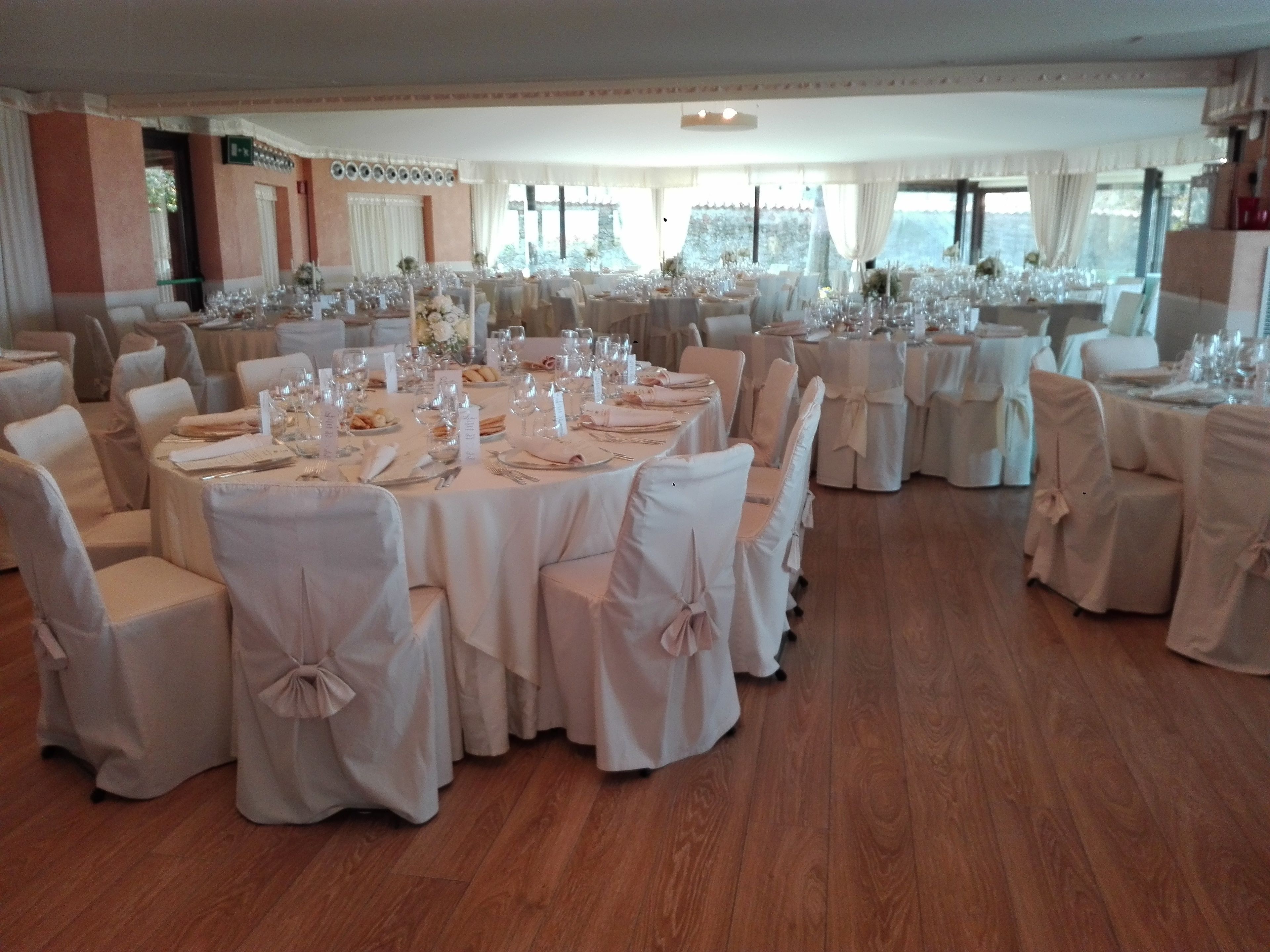 Un salón de banquetes con mesas redondas dispuestas para comer, sillas con fundas blancas y suelo de madera.