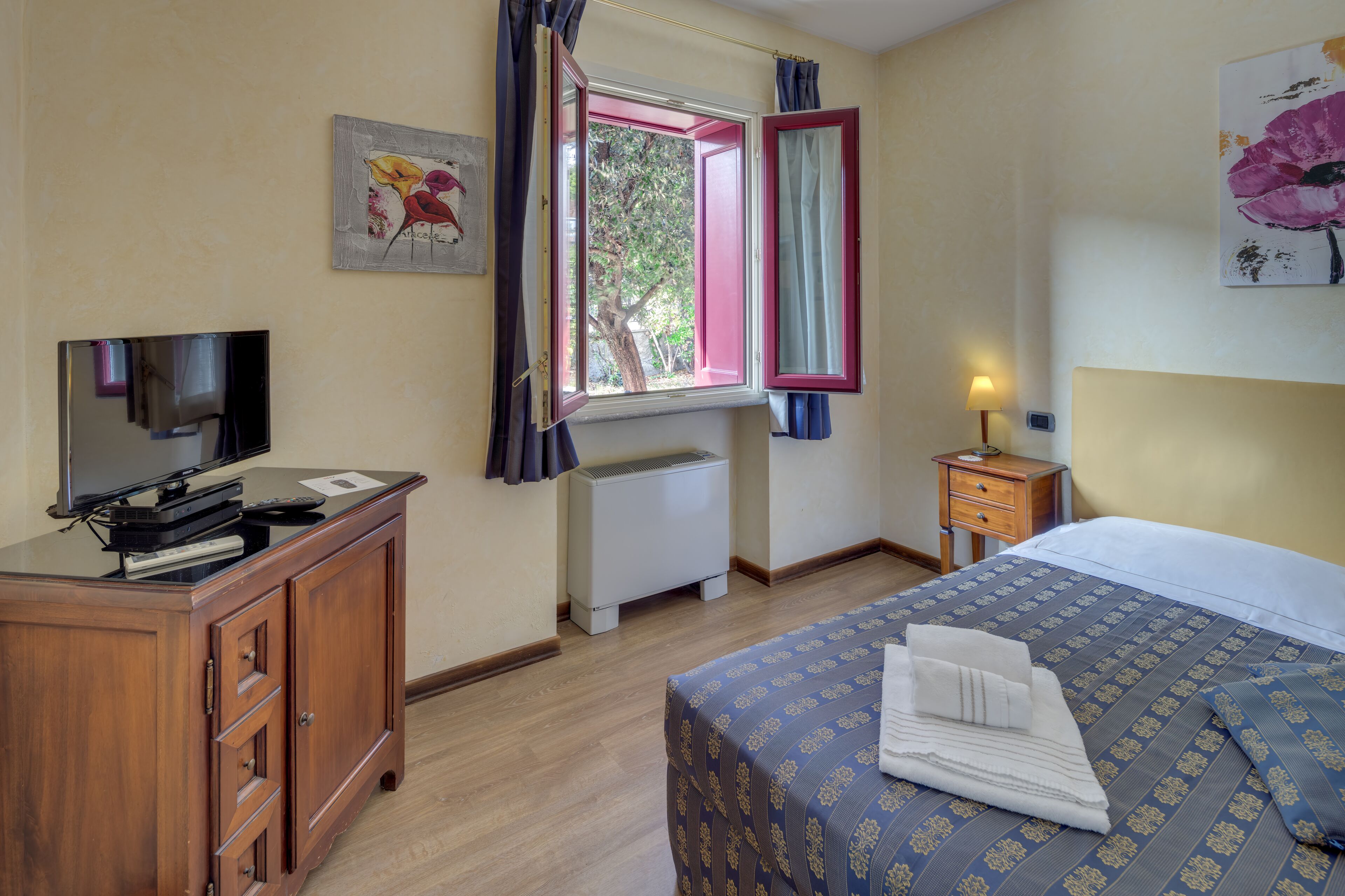 A hotel room with a bed, a desk with a television, a window with curtains, and a nightstand with a lamp.