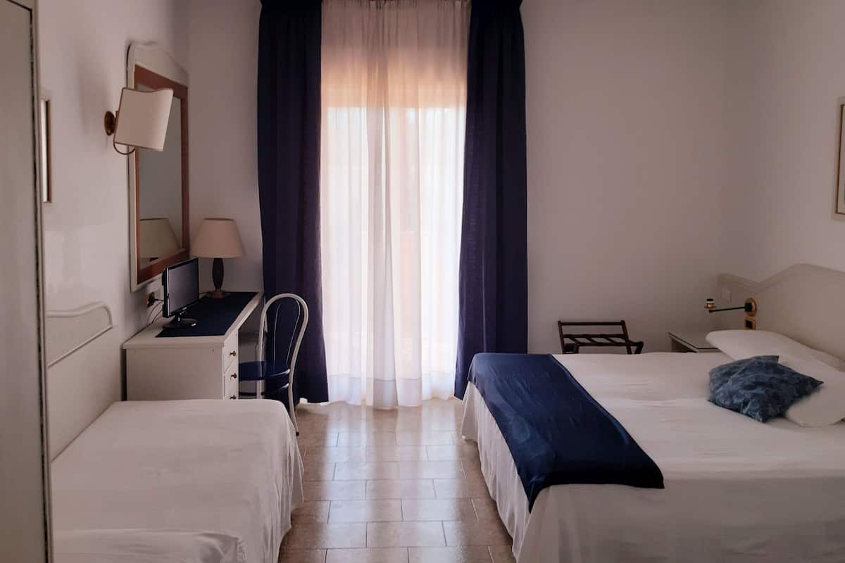 A hotel room with two beds, a desk, a chair, a lamp, a mirror, and a window with curtains.