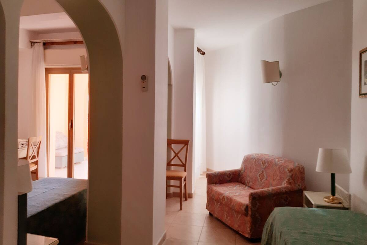 A hotel room with a bed, a chair, a small table, and a lamp. There is a door leading to a balcony.