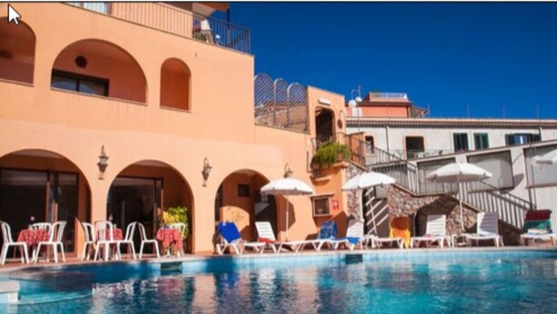 A poolside area with lounge chairs and umbrellas, a clear blue pool, and a building with arched windows and balconies.