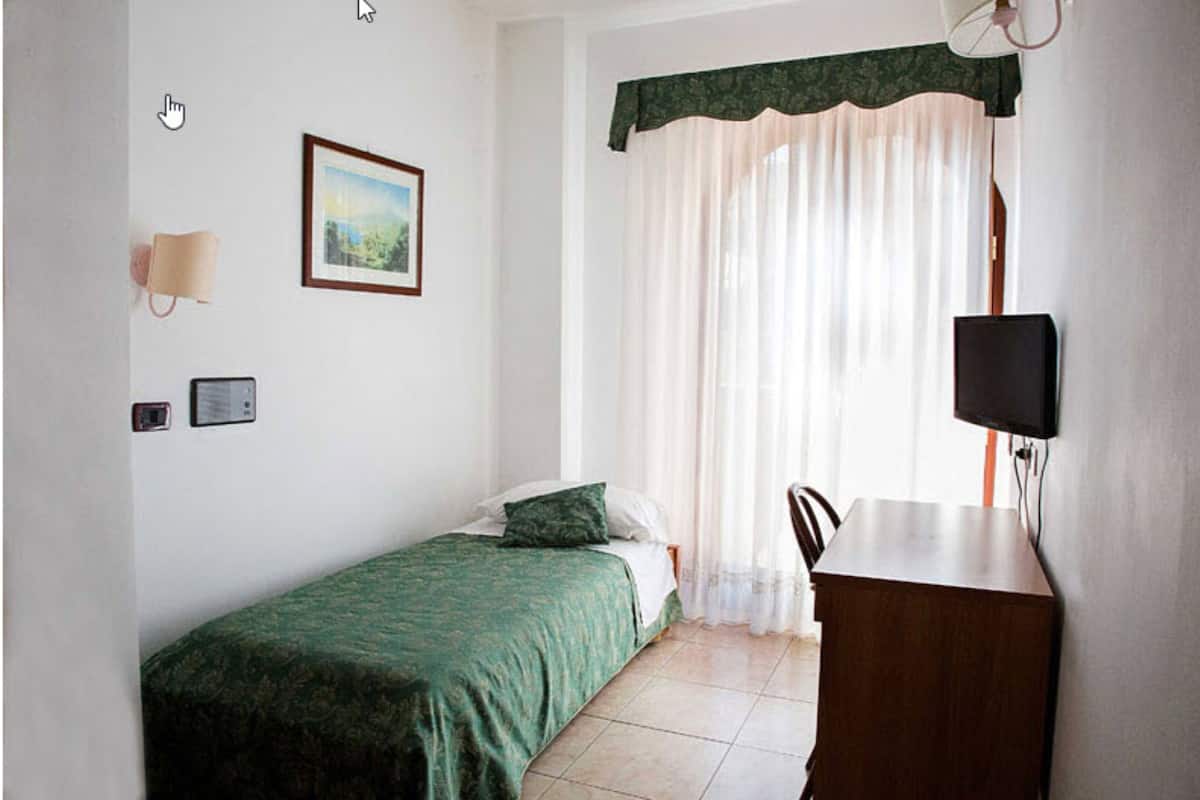 A small, simple room with a bed, a desk, a chair, a lamp, and a window with curtains.