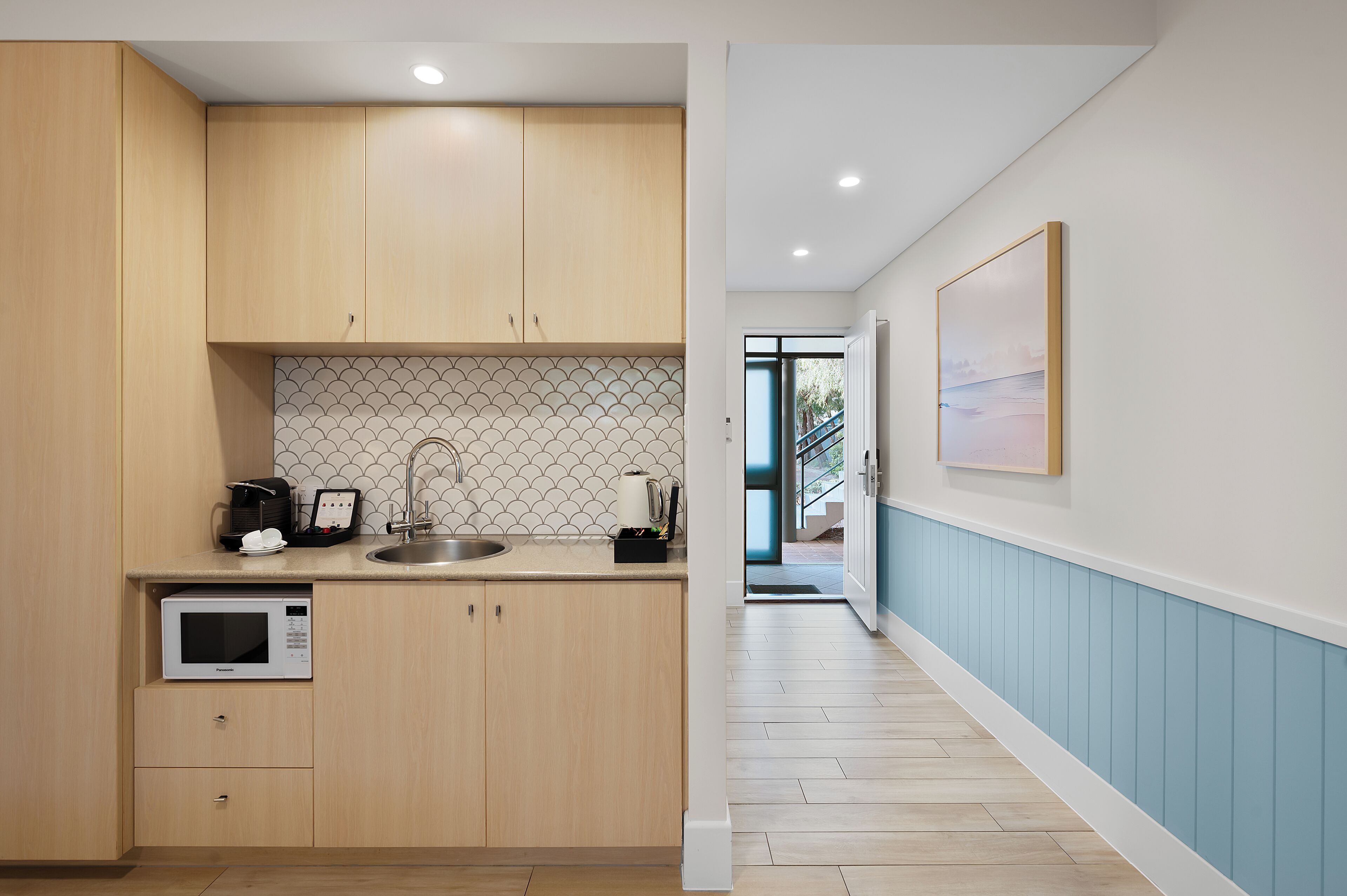 A modern kitchen with wooden cabinets, a microwave, and a sink. There is a hallway with blue walls and a door leading outside.