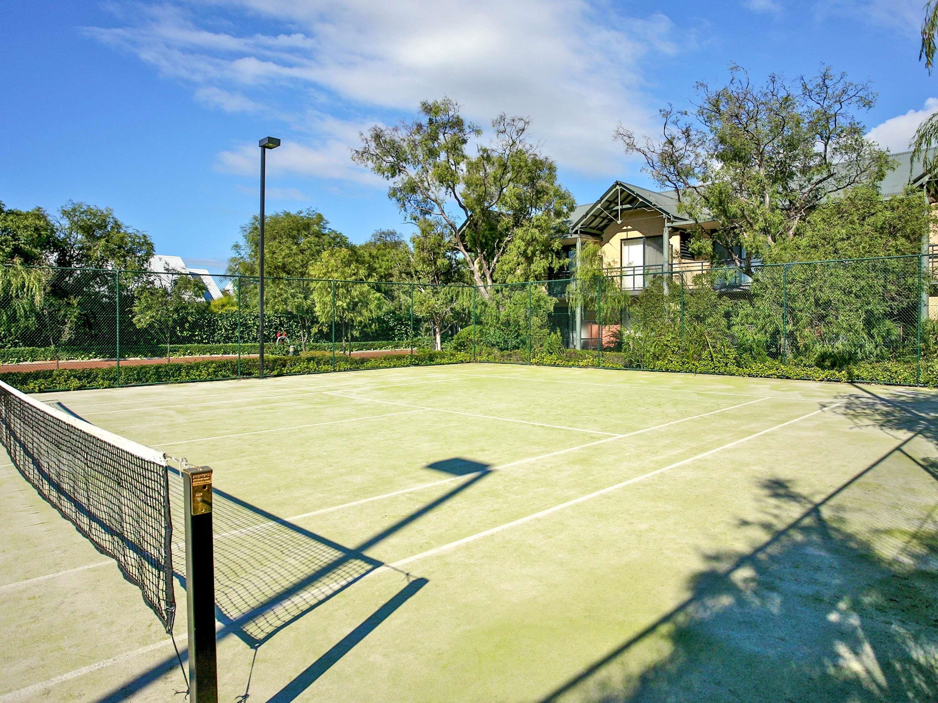 Ein Tennisplatz im Freien mit Netz, umgeben von Bäumen und einem Gebäude im Hintergrund.