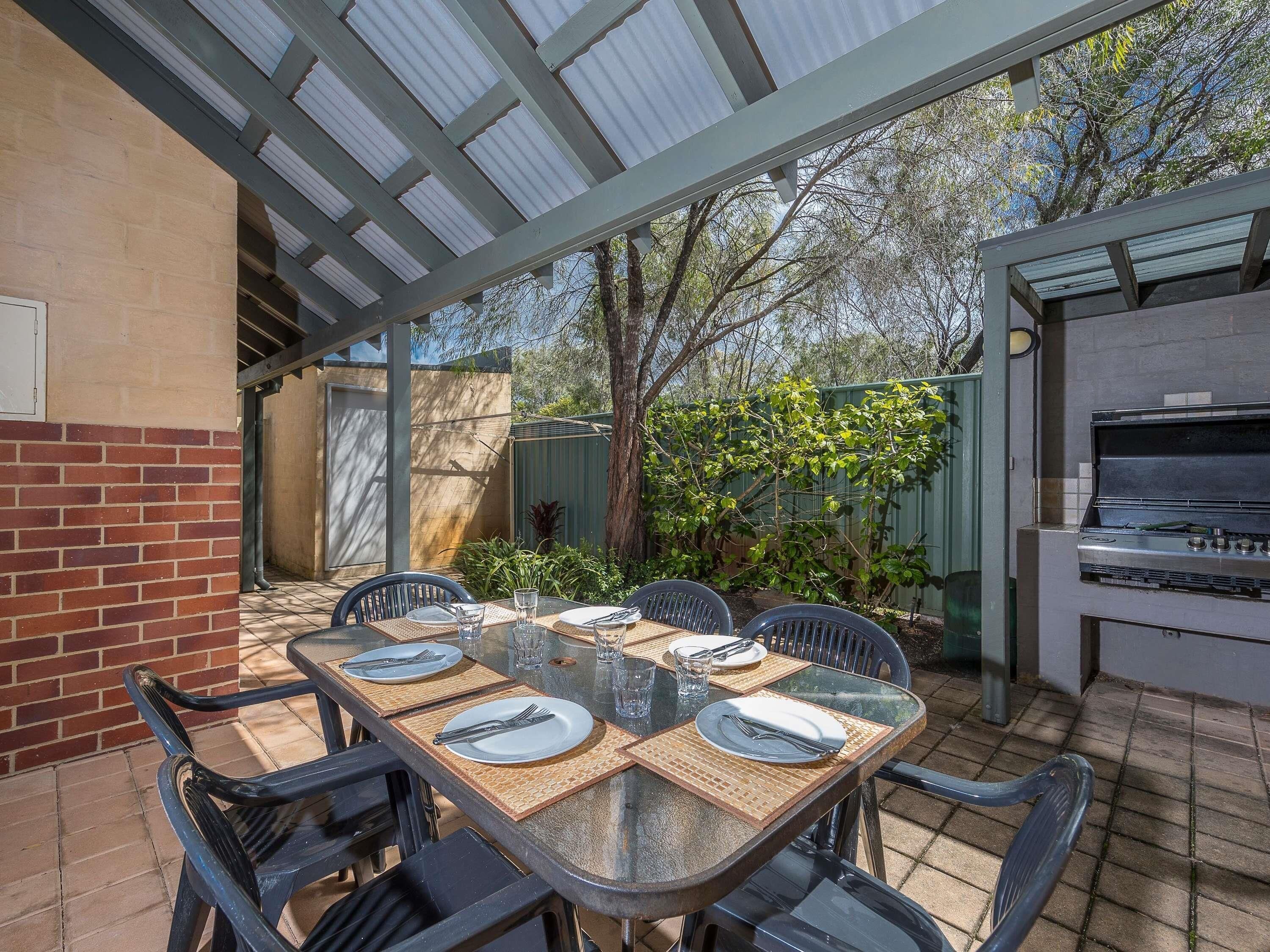 Une terrasse extérieure avec une table en verre pour quatre personnes, entourée de chaises, d’un barbecue et d’un mur en briques.