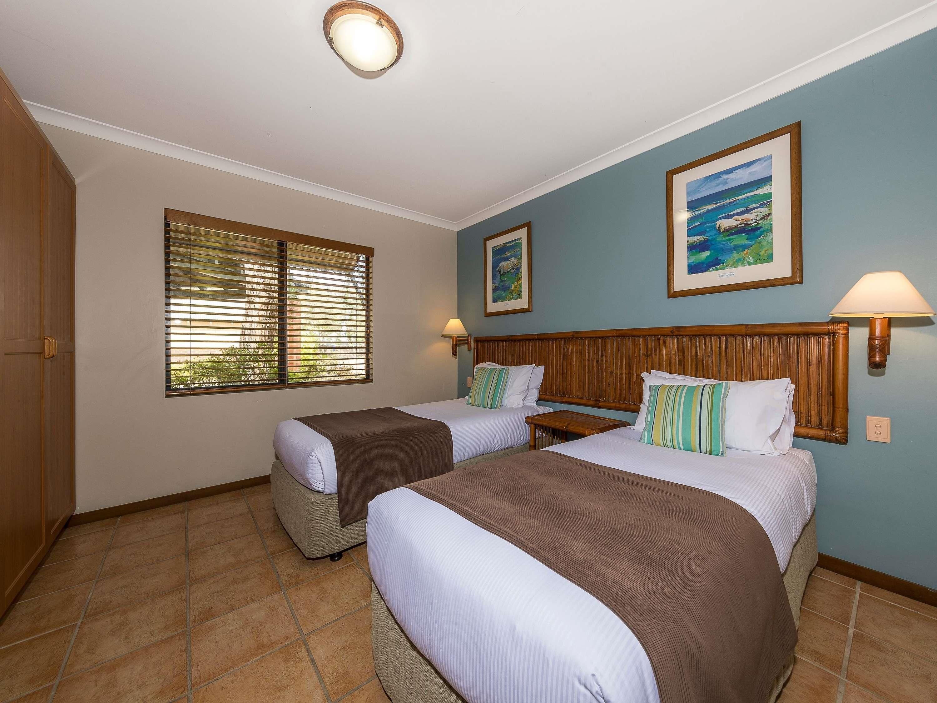 Quarto de hotel com duas camas, uma janela com persianas, um quadro na parede e uma luminária.