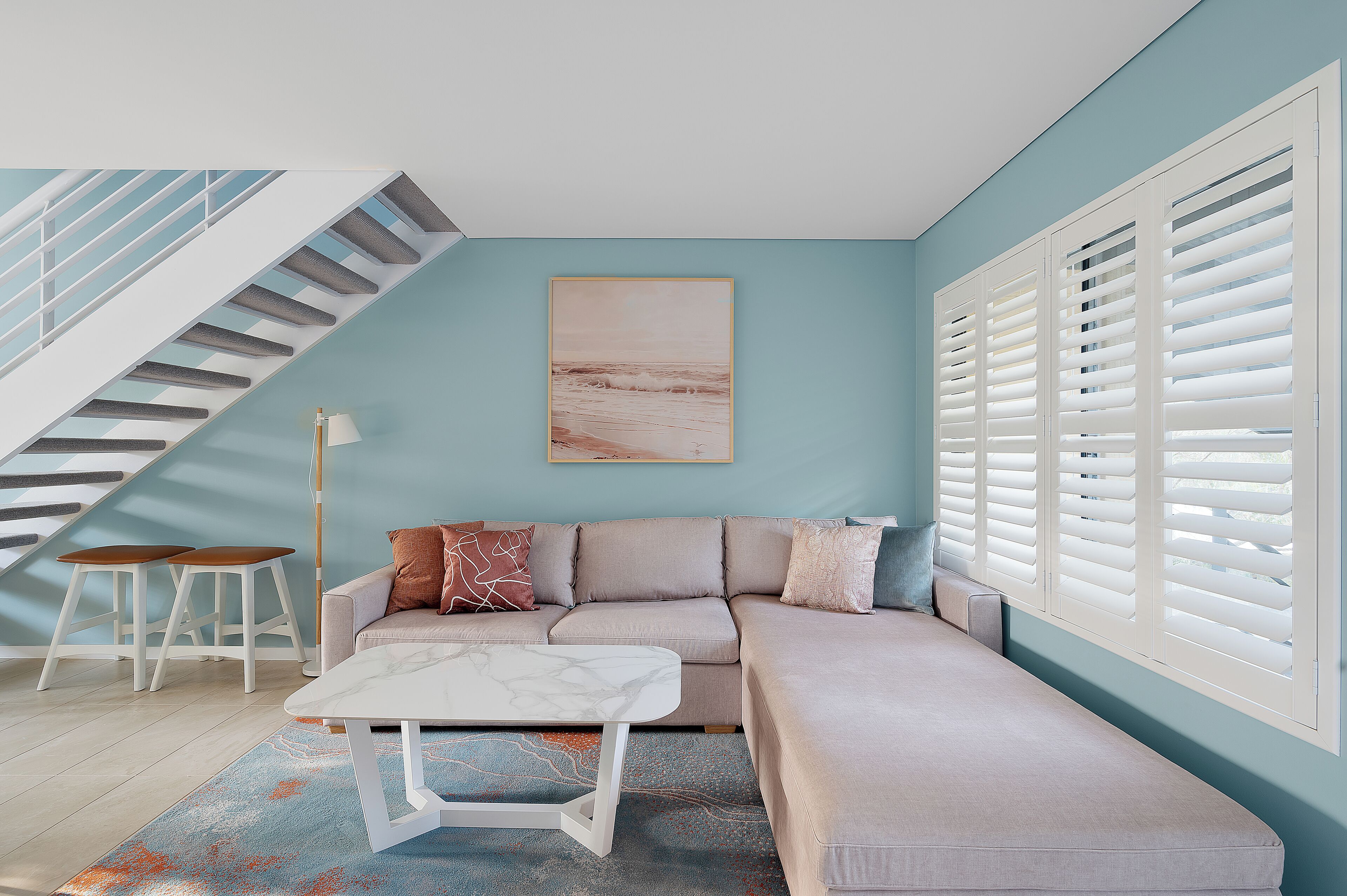 A modern living room with a staircase, a sofa, a coffee table, and a large window with shutters.