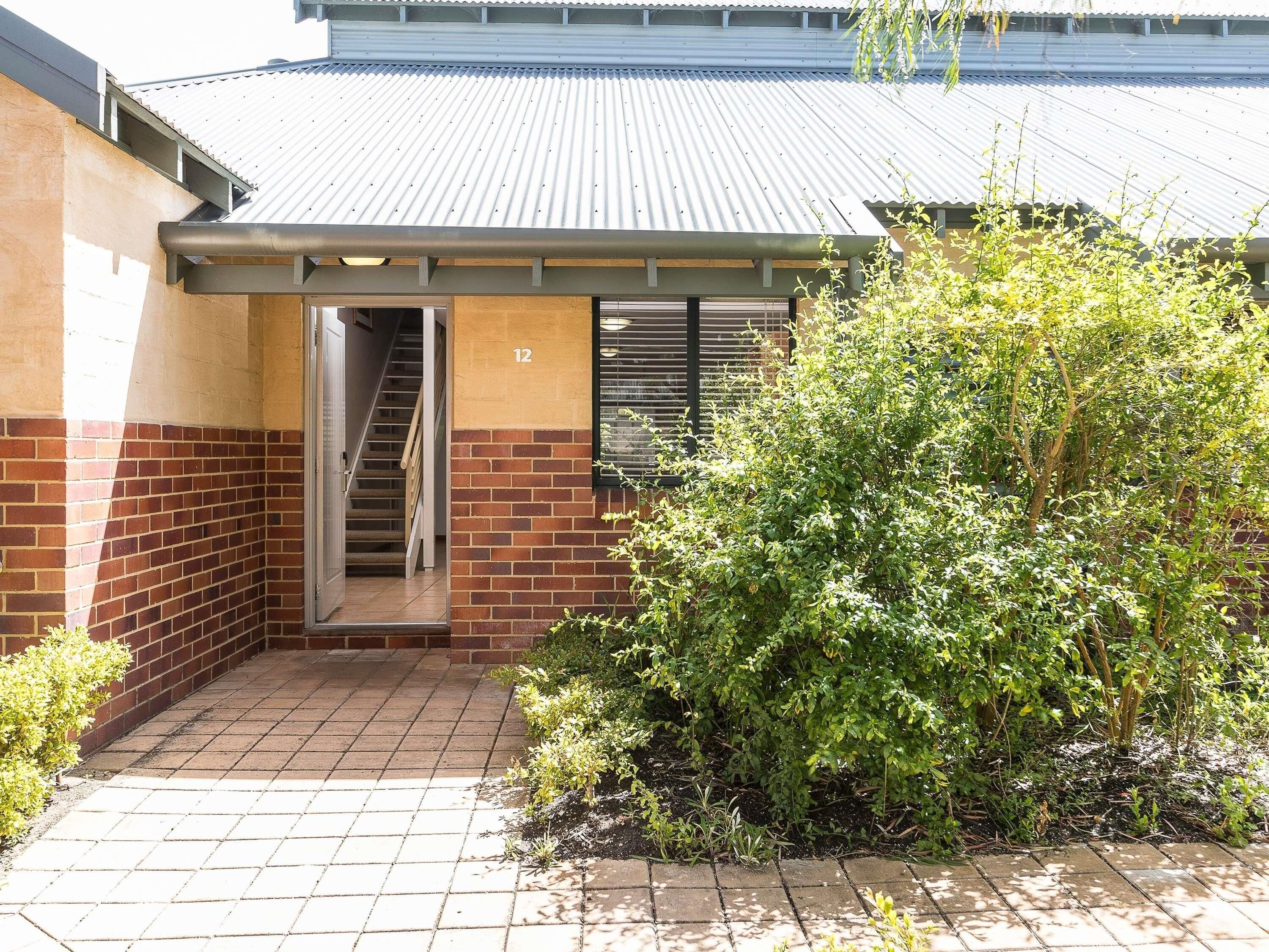 A building with a brick facade, a metal roof, a staircase leading to an entrance, and a small garden area with shrubs.