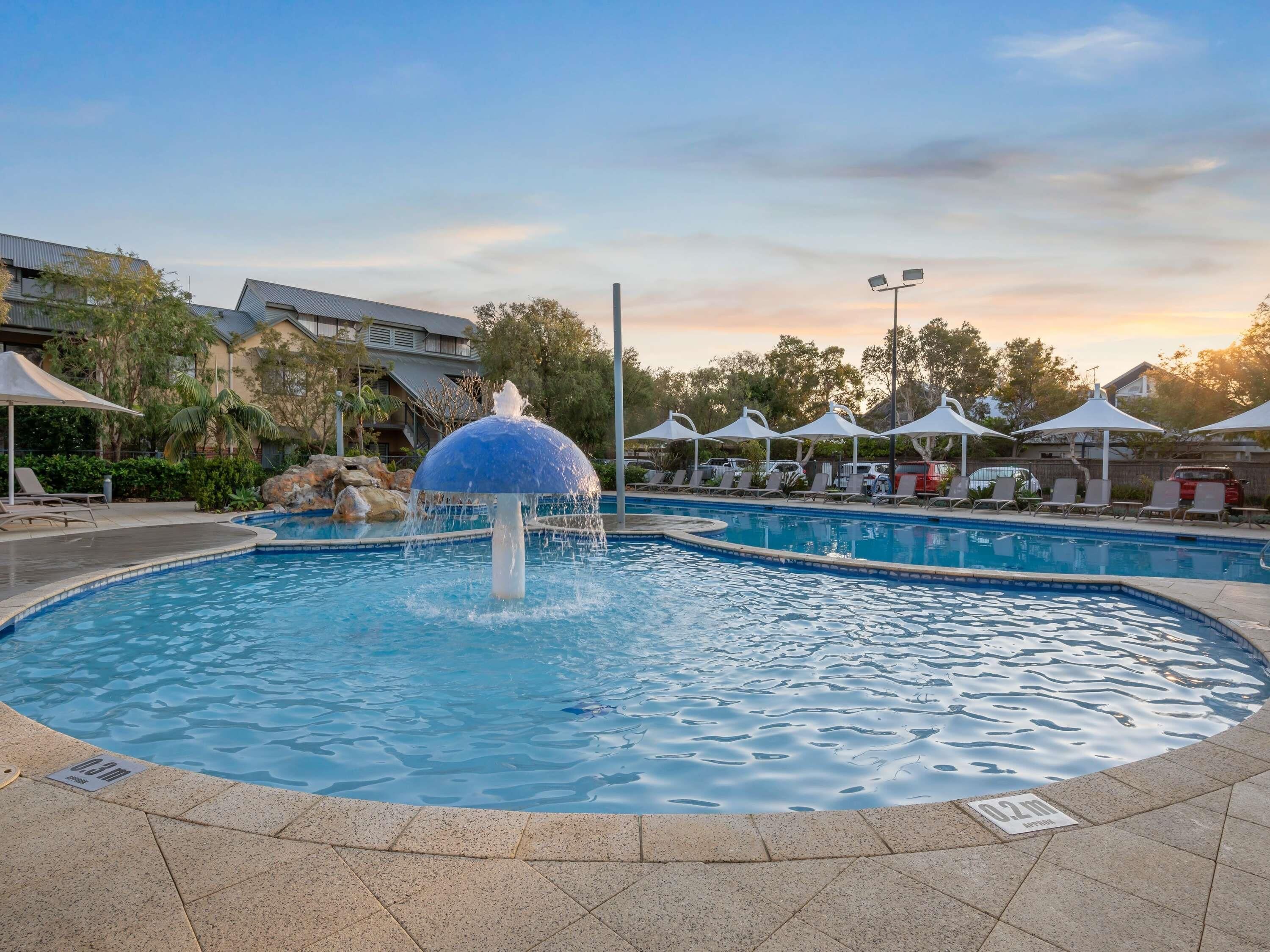 A swimming pool with a water fountain, lounge chairs, umbrellas, and a building in the background.