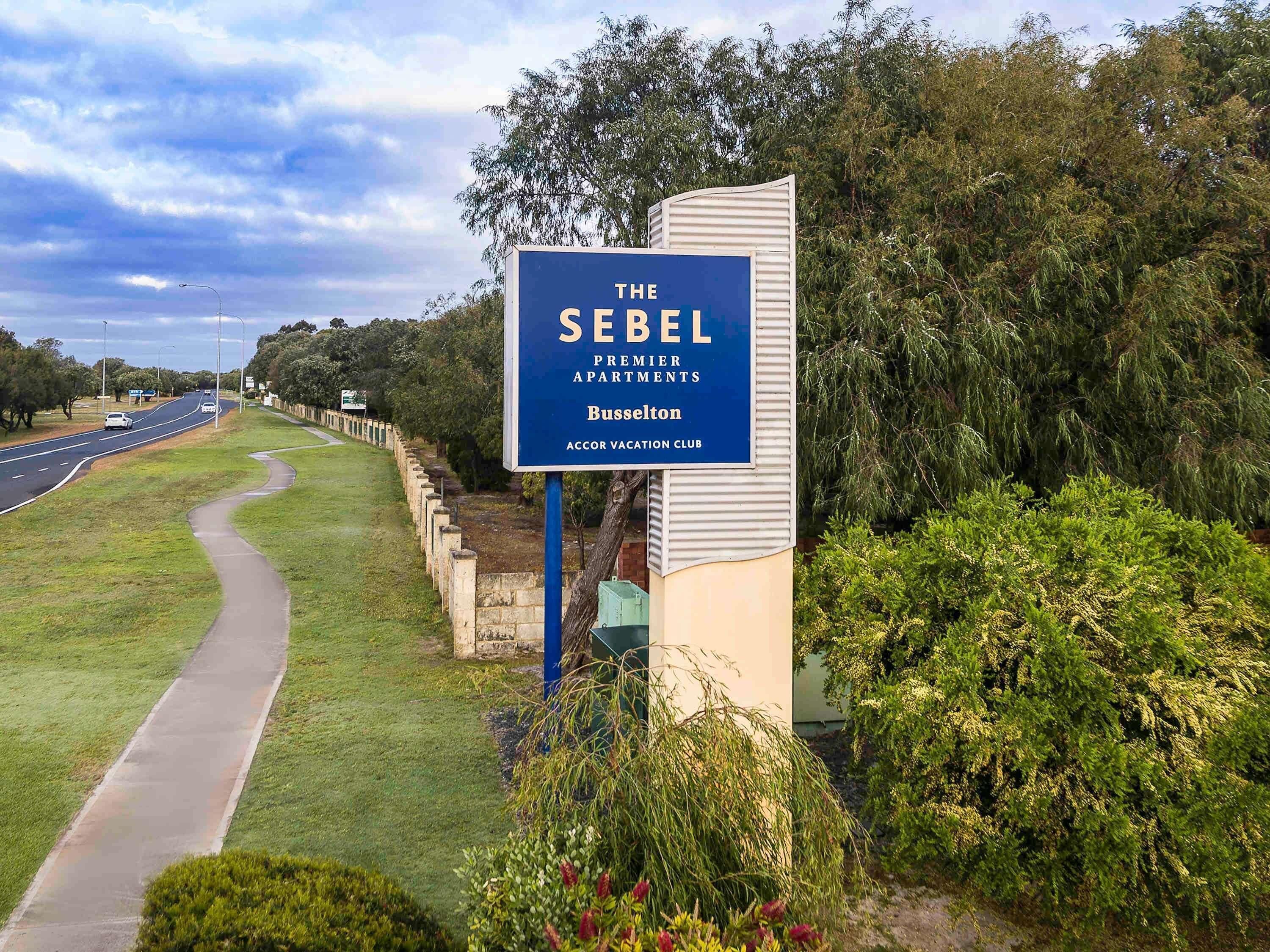 Placa indicando The Sebel Premier Apartments em Busselton, com um caminho e vegetação ao fundo.