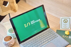 Una computadora portátil con una pantalla verde mostrando el logotipo de EXO BOUTIQUE HOTEL, una escultura de madera, dos cuencos decorativos, un patito de goma y un letrero de prohibición de fumar.
