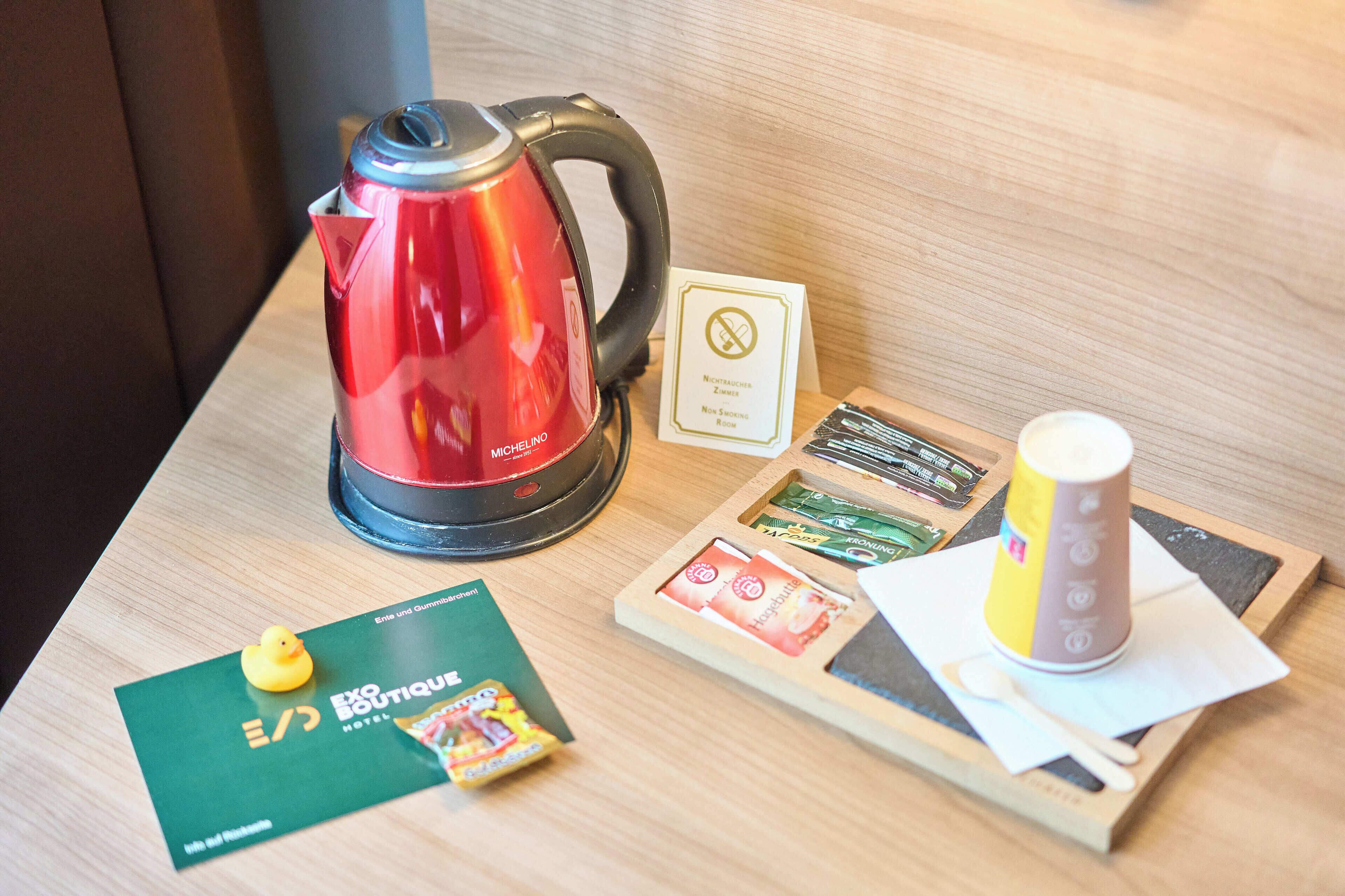 Una habitación de hotel con un hervidor rojo, una tarjeta con el letrero de no fumar, una tarjeta con el logotipo del hotel, una bandeja con una taza y una cuchara, y un pequeño patito amarillo de juguete.