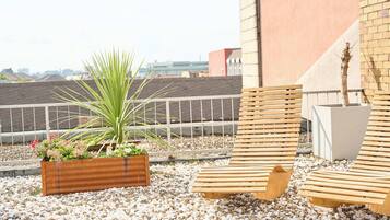 Eine Dachterrasse mit Holzliegen, einem Pflanzgefäß mit Pflanzen und Blick auf Gebäude im Hintergrund.