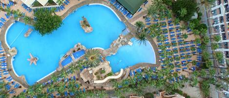 An aerial view of a resort with a large central pool, multiple smaller pools, and numerous sun loungers arranged around the pool area.