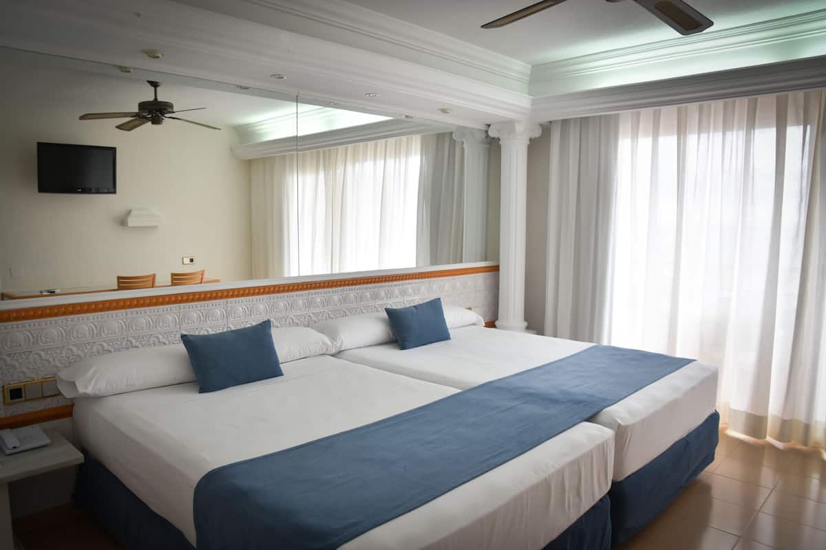 A hotel room with two beds, a ceiling fan, a flat-screen TV, and a large window with sheer curtains.