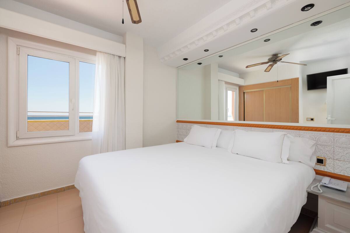 A hotel room with a large bed, a ceiling fan, a mirror, and a window with curtains.