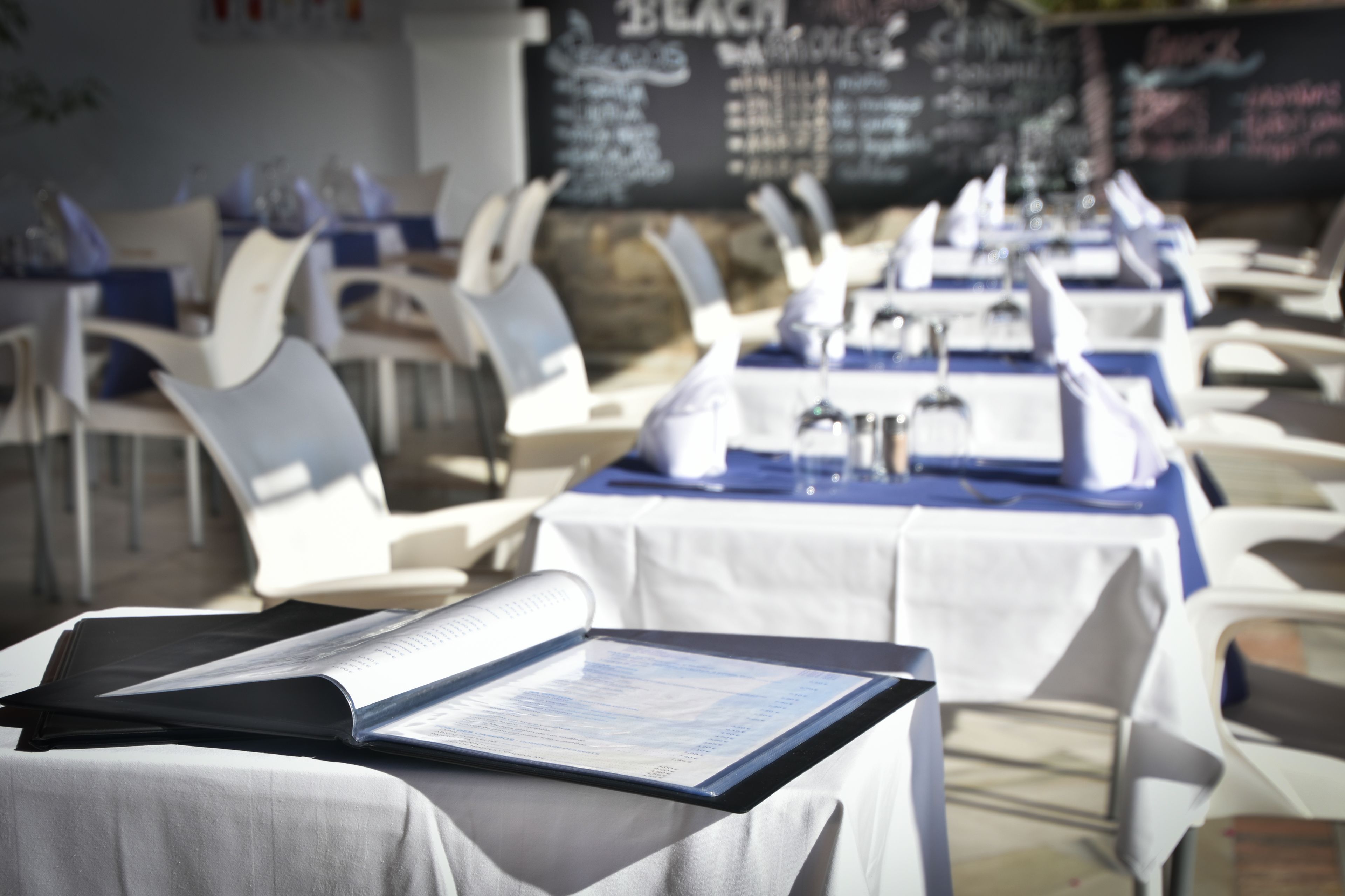 Ein Restaurant mit weißen Stühlen und Tischen, Speisekarten auf den Tischen und einer Kreidetafel mit der Speisekarte im Hintergrund.