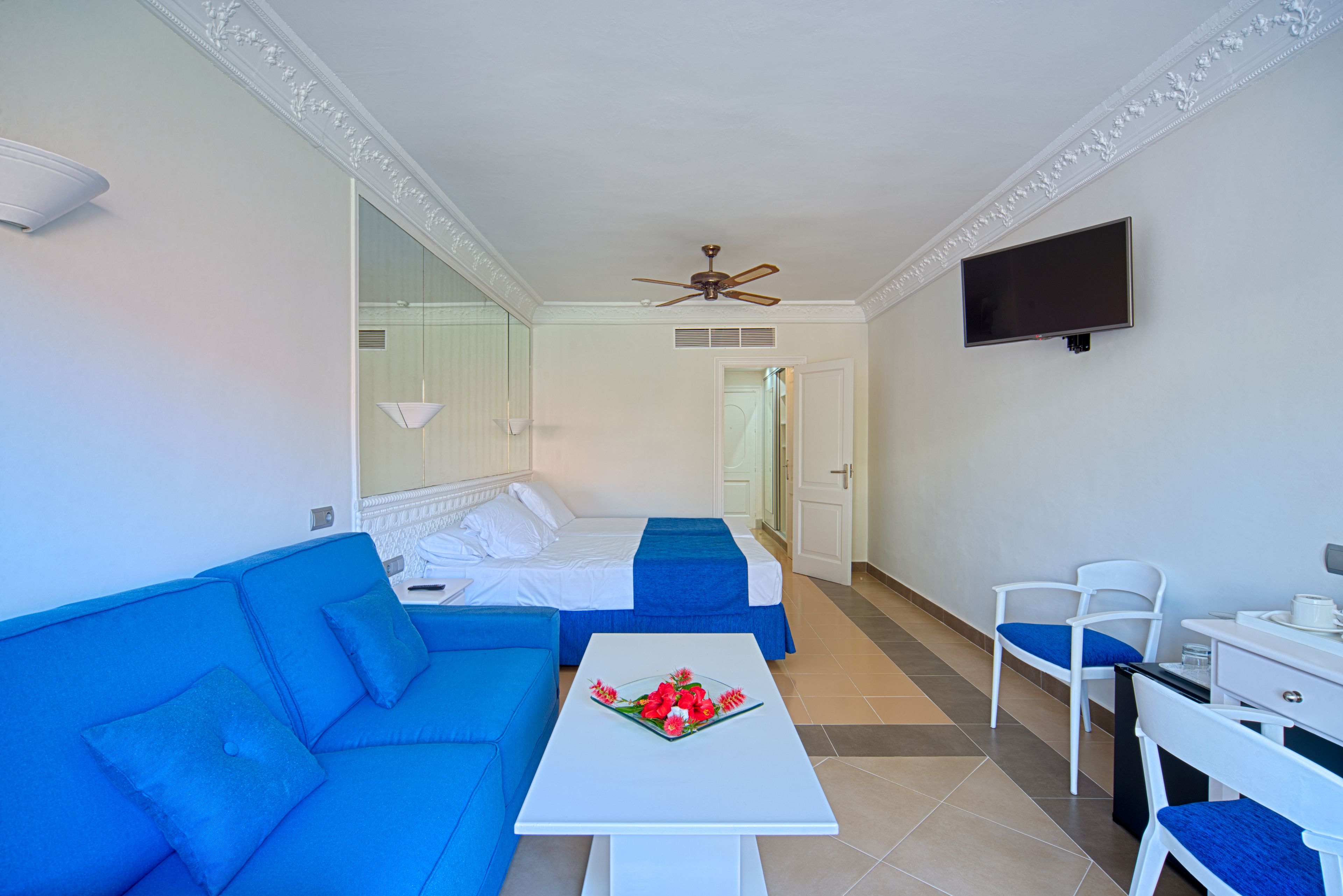 Habitación de hotel con sofá azul, cama blanca con cubrecama azul, mesa blanca con un centro de mesa floral, escritorio blanco con una silla y un ventilador de techo.