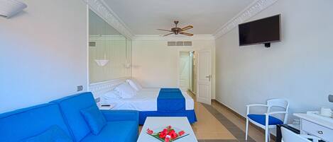 Habitación de hotel con sofá azul, cama blanca con cubrecama azul, mesa blanca con un centro de mesa floral, escritorio blanco con una silla y un ventilador de techo.