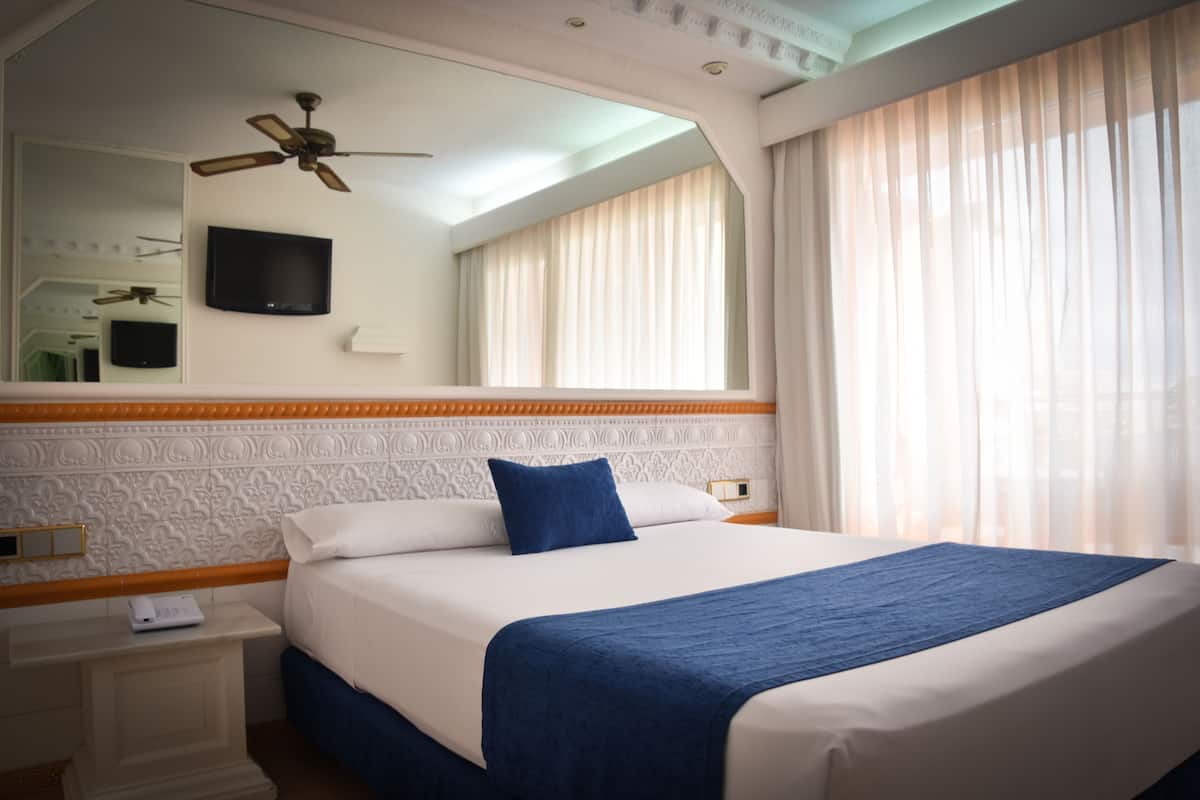A hotel room with a large bed, a ceiling fan, a flat-screen TV, and a window with sheer curtains.