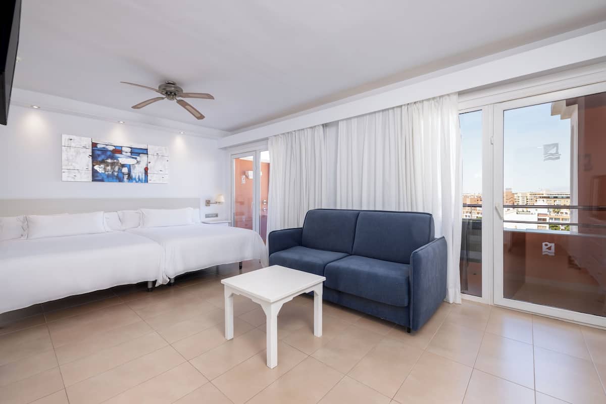 A modern hotel room with a white sofa, a blue sofa, a small white table, a ceiling fan, a large window with curtains, and a city view.