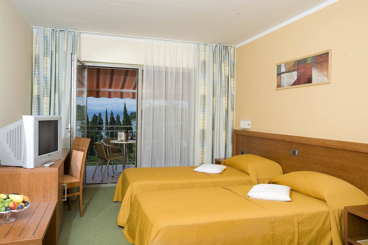 A hotel room with two beds, a desk with a television, and a balcony with a view of trees and a building.