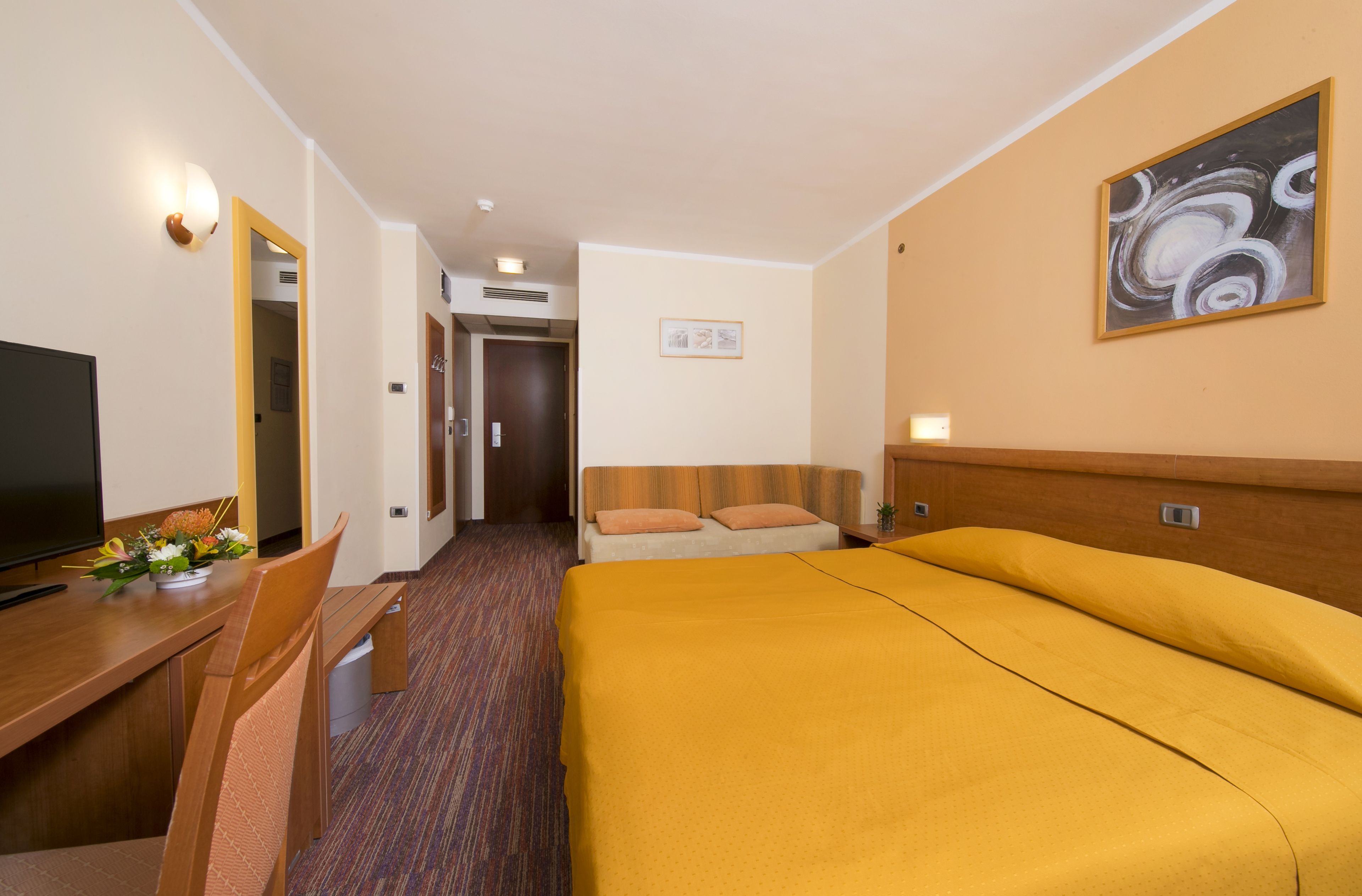Habitació d'hotel amb un llit, una escriptori amb televisió, una cadira i una petita taula amb flors.