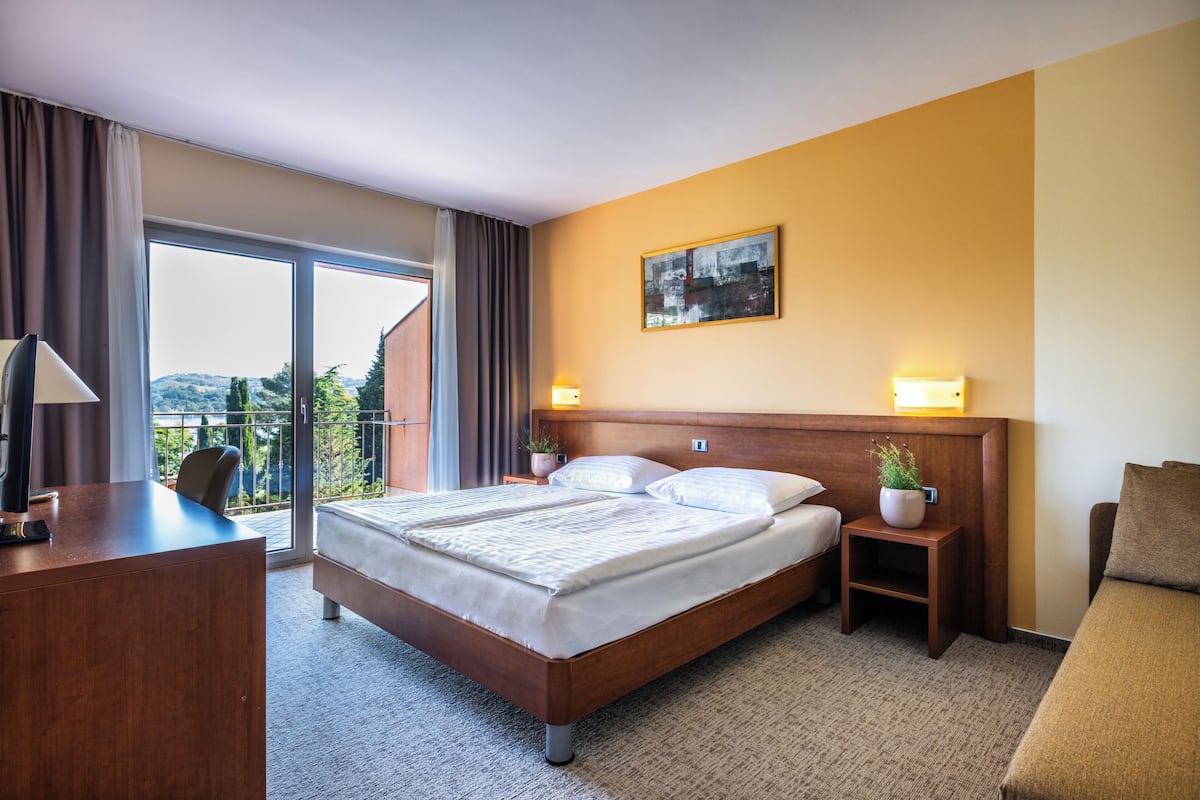 A hotel room with a bed, desk, chair, bedside tables, and a balcony with curtains.