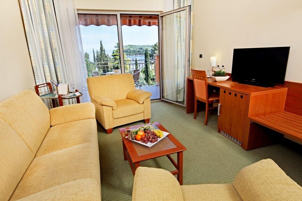 Ein Hotelzimmer mit einer Couch, einem Sessel, einem kleinen Tisch mit Obst, einem Fernseher und einem Balkon mit Aussicht.