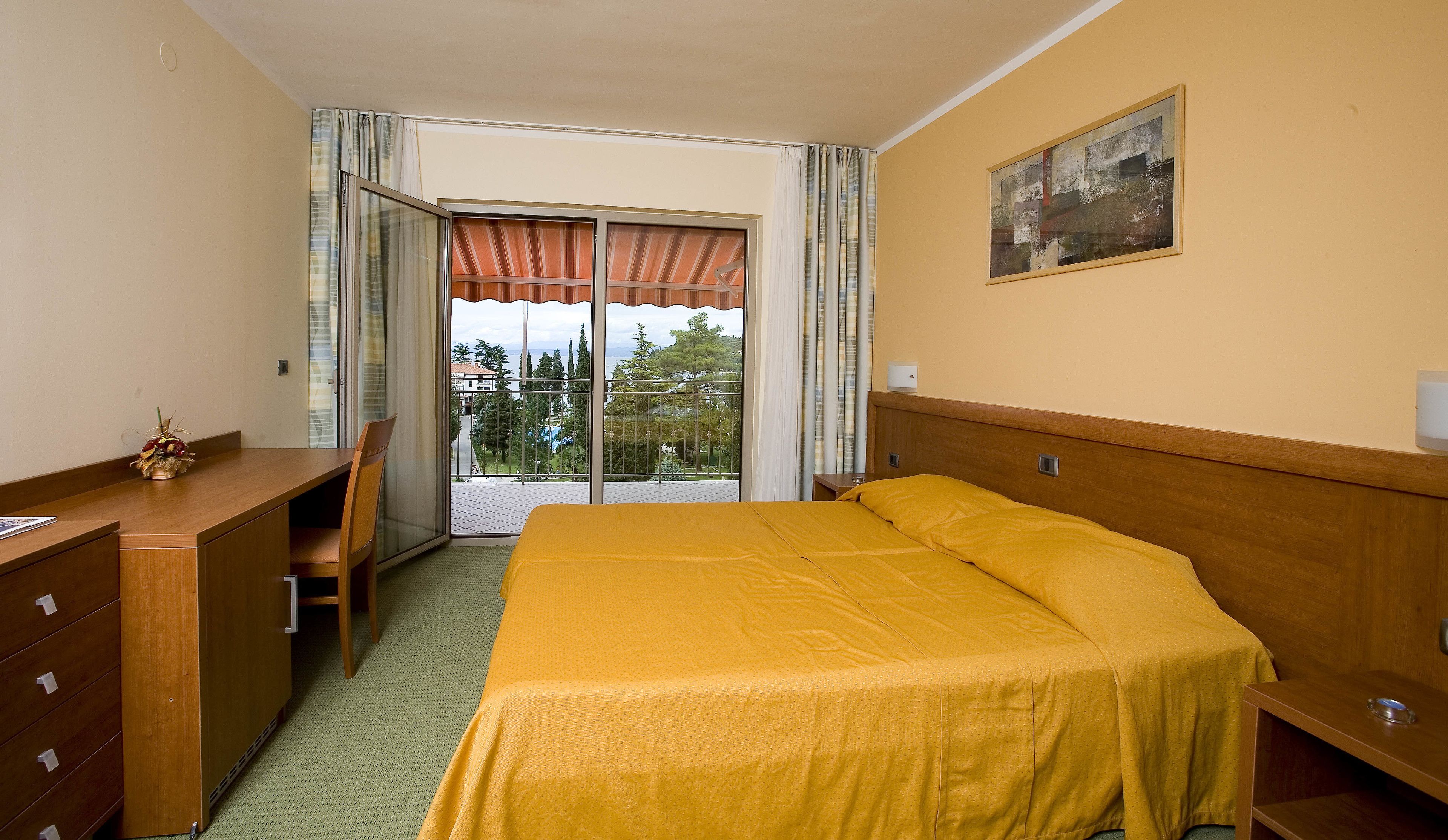 A hotel room with a bed, a desk, and a chair. There is a balcony with a view of trees and buildings.