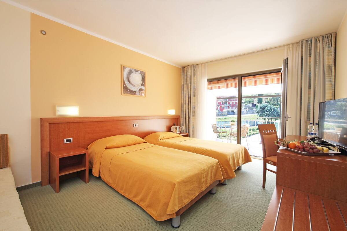 A hotel room with two beds, a desk with a TV, and a balcony with a view of buildings.