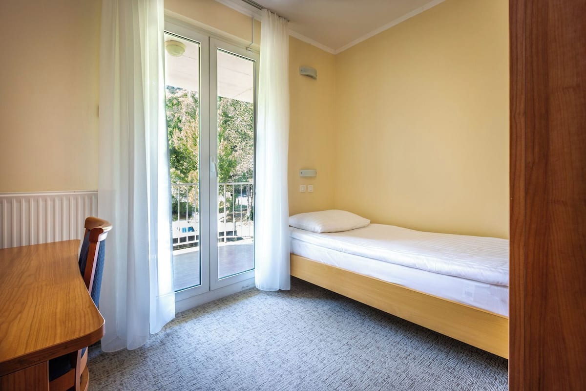 A small room with a single bed, a wooden table, and a sliding glass door leading to an outdoor area.