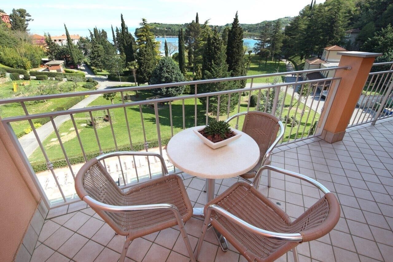 Un balcón con sillas de mimbre y una mesa, con vista a un jardín ajardinado y una masa de agua.