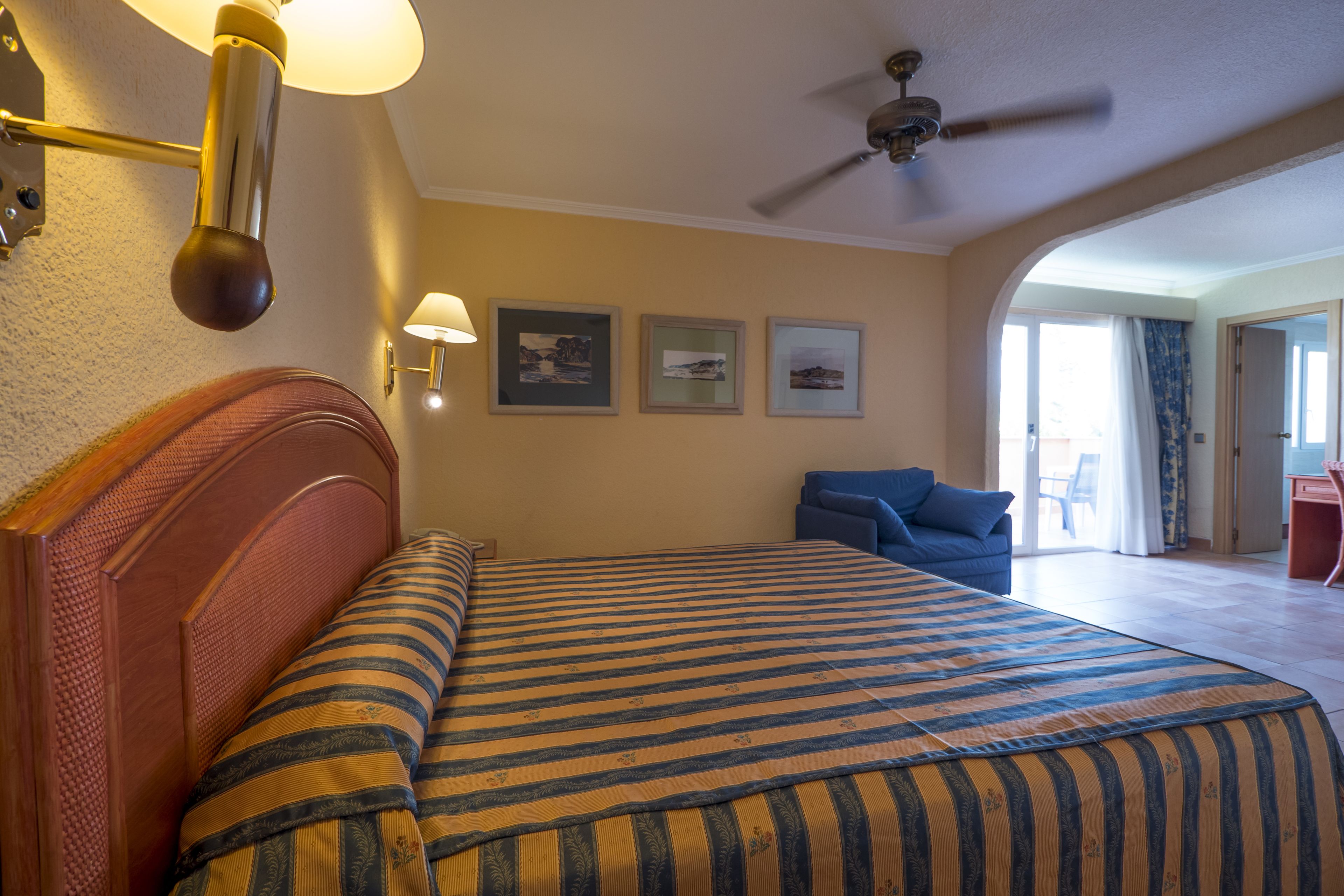 Habitación de hotel con cama, ventilador de techo, escritorio y vistas al exterior.
