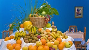 Una mesa con varias frutas y una cesta de mimbre sobre un mantel, frente a una pared azul con un cuadro enmarcado.