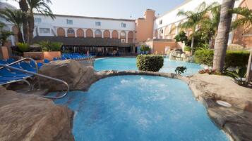 Zona de piscina con sillas reclinables azules, borde de piedra y un edificio con ventanas arqueadas al fondo.